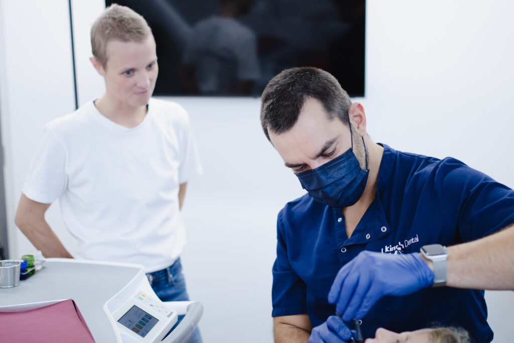 Zahnarzt mit Mundschutz und Handschuhen behandelt einen Patienten, ein Kollege schaut zu